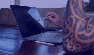 A tattooed person using a stylus to point at a laptop screen on a wooden table, reviewing digital content.